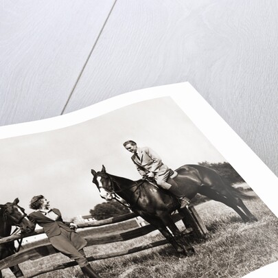 1930s 1940s couple in riding gear man riding horse woman standing by wooden fence by Anonymous