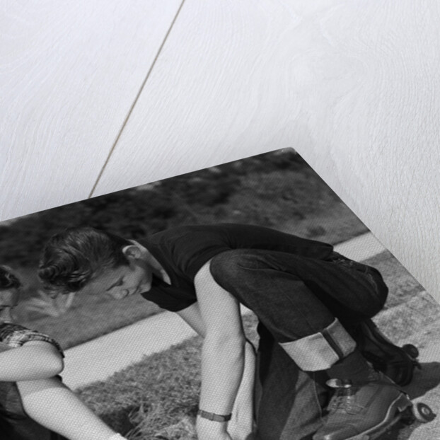 1950s teen boy helping girl put on metal roller skates sitting on sidewalk by Anonymous