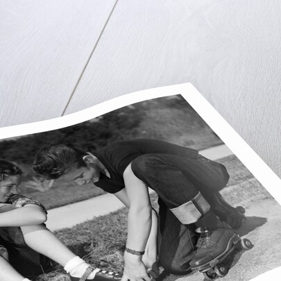 1950s teen boy helping girl put on metal roller skates sitting on sidewalk by Anonymous