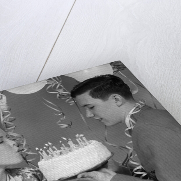 1960s teenage girl blowing out candles on birthday cake held by boy by Anonymous