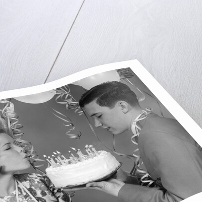 1960s teenage girl blowing out candles on birthday cake held by boy by Anonymous