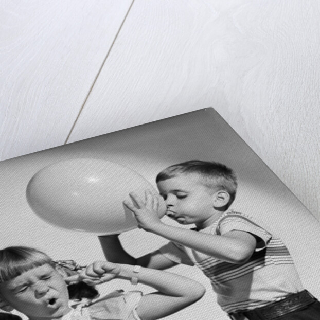 1950s little boy blowing up big balloon little girl with fingers in ears eyes closed by Anonymous
