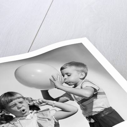 1950s little boy blowing up big balloon little girl with fingers in ears eyes closed by Anonymous