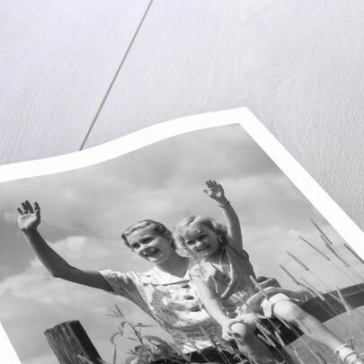 1930s girl sitting on fence with woman next to her in field waving by Anonymous