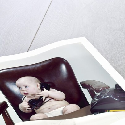 1960s chubby baby sitting in leather chair at office desk holding telephone by Anonymous