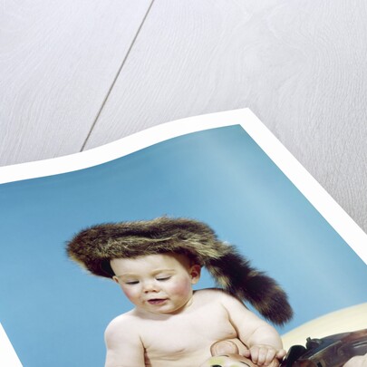 1960s baby boy wearing coonskin cap with stuffed animal and shotgun by Anonymous