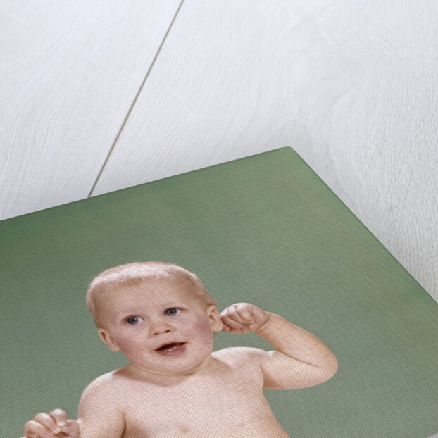 1960s baby wearing diaper sitting on pink blanket mouth open hand touching ear by Anonymous