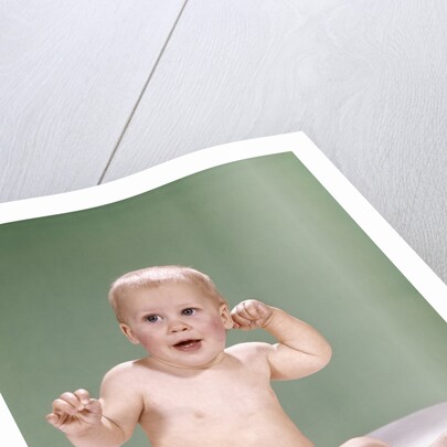 1960s baby wearing diaper sitting on pink blanket mouth open hand touching ear by Anonymous