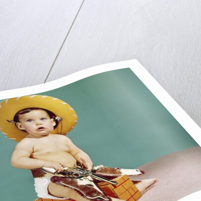 1960s baby wearing cowboy hat and holster with guns pistols looking at camera by Anonymous