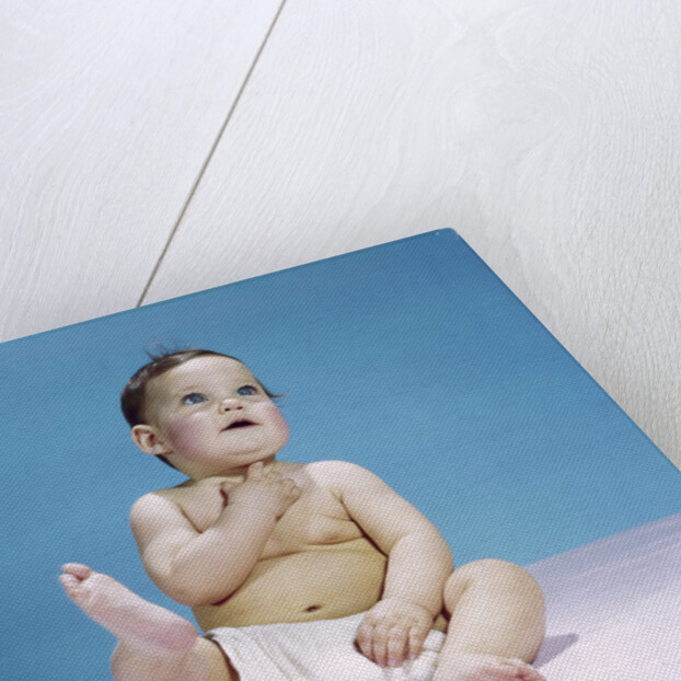 1960s baby wearing diaper sitting looking up and kicking one leg up by Anonymous