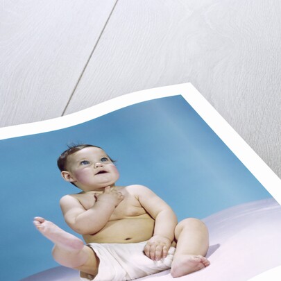 1960s baby wearing diaper sitting looking up and kicking one leg up by Anonymous