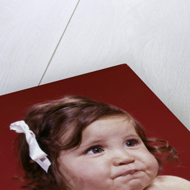 1960s portrait of chubby unhappy baby girl with white ribbon bow in messy hair by Anonymous