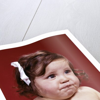 1960s portrait of chubby unhappy baby girl with white ribbon bow in messy hair by Anonymous