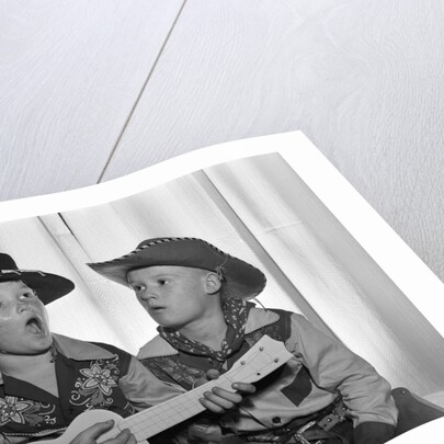 1950s 2 juvenile boys in cowboy hat & shirts playing ukulele & singing mouth open wide by Anonymous