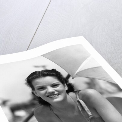 1930s teen girl lying on beach under umbrella wearing bathing suit smiling looking at camera by Anonymous