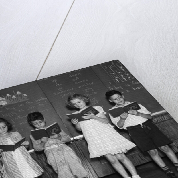 1930s line-up of 5 elementary school students in front of blackboard reading books with teacher looking on by Anonymous
