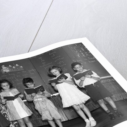1930s line-up of 5 elementary school students in front of blackboard reading books with teacher looking on by Anonymous