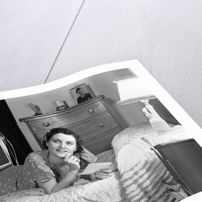 1940s fully dressed smiling woman laying on her bed with her legs up reading a letter in her bedroom by Anonymous