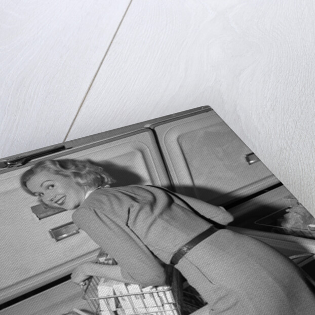 1950s blond woman lifting wire basket food items from a deep freezer looking at camera over her shoulder by Anonymous