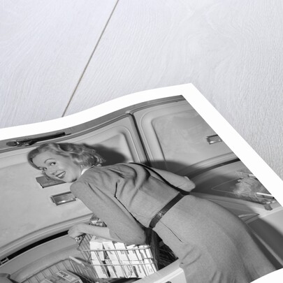 1950s blond woman lifting wire basket food items from a deep freezer looking at camera over her shoulder by Anonymous