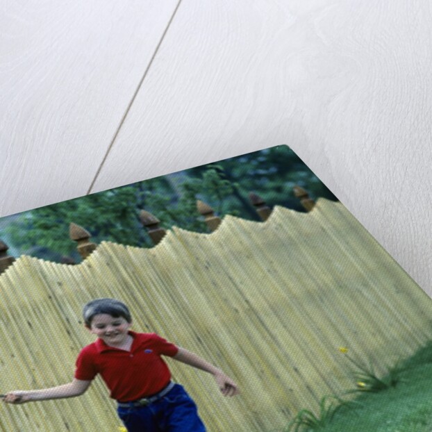 1980s Smiling Boy Running Along Sidewalk Rattling Stick On Tall Picket Fence Making Noise by Anonymous