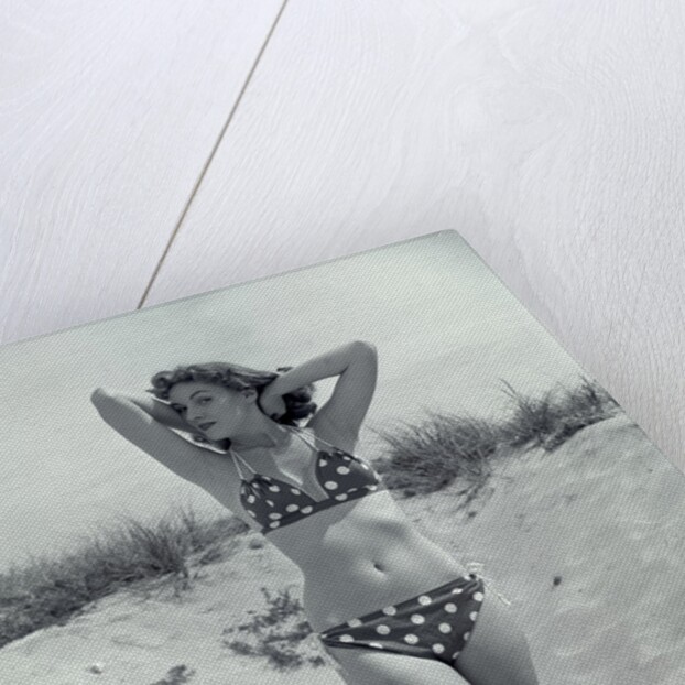 1950s Brunette Bathing Beauty In Polka Dot Bikini Standing In Sand With Hands Behind Head Looking At Camera by Anonymous