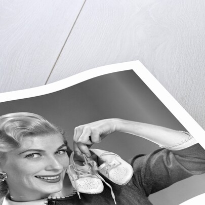 1950s 1960s Smiling Blond Woman Communicating That She Is Mother To Be By Holding Up A Pair Of Baby Shoes Looking At Camera by Anonymous