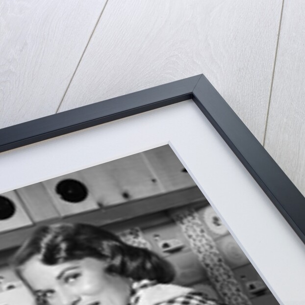1950s Housewife Wearing Checkered Dress Standing In Kitchen Stirring Pot On Stove Looking Over Shoulder by Anonymous