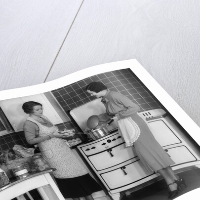 1930s Woman Housewife And Friend Wearing Apron Cooking Food In Kitchen On Gas Stove Indoor by Anonymous