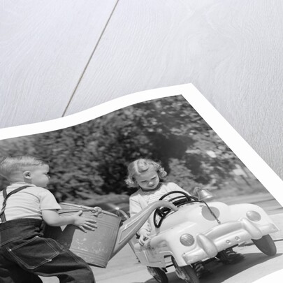 1950s Little Boy Playing Gas Station Pouring Water Into Toy Car For Little Girl by Anonymous