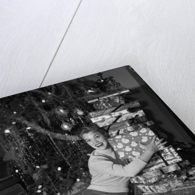 1950s portrait of young woman holding stack of presents next to a Christmas tree by Anonymous