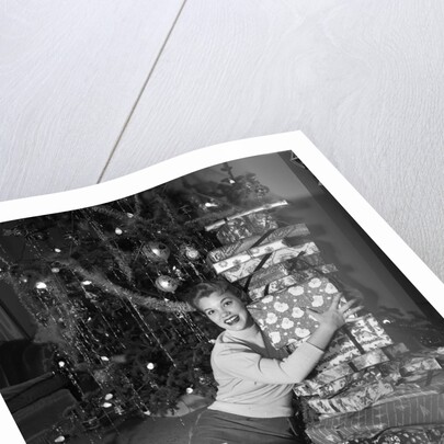 1950s portrait of young woman holding stack of presents next to a Christmas tree by Anonymous