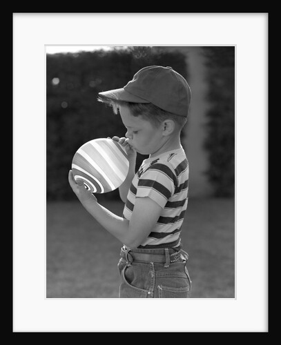1950s Side View Of Boy In Baseball Cap and Striped T-Shirt Blowing Up Swirled Balloon by Anonymous