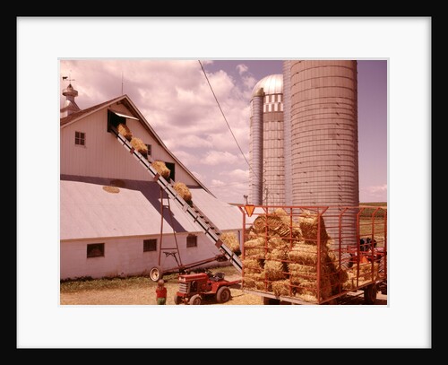 1970s Girl Watching Hay Bales On Conveyor Belt Loading Into Barn By Farm Grain Silos by Anonymous