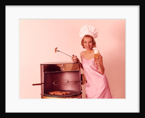 1960s Woman Wearing Pink Party Dress And Chef Hat Grilling Hot Dogs Drinking Beer by Anonymous