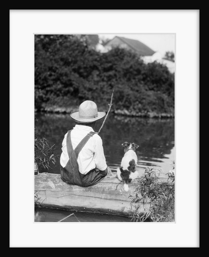 1920s 1930s Farm Boy Wearing Straw Hat And Overalls Sitting On Log With Spotted Dog Fishing In Pond by Anonymous