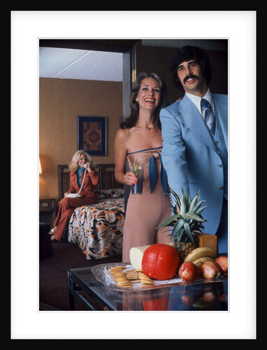 1970s Couple Welcoming Guest To Party At Home Woman Using Phone In Another Room by Anonymous