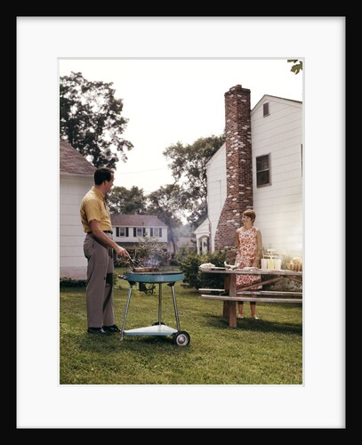 1960s Couple Suburban Backyard Man Grilling Woman Setting Picnic Table by Anonymous
