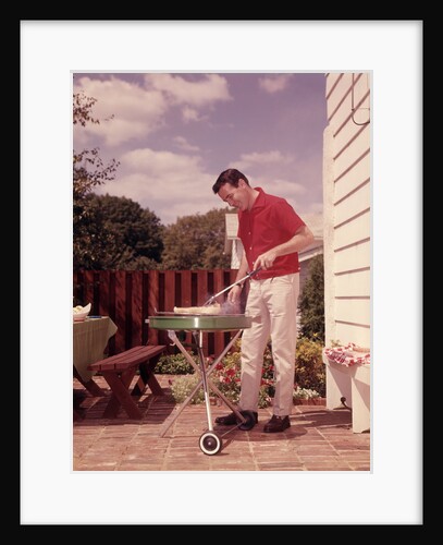 1960s Man Wearing Red Shirt Cooking Steak Outdoor On Backyard Grill by Anonymous