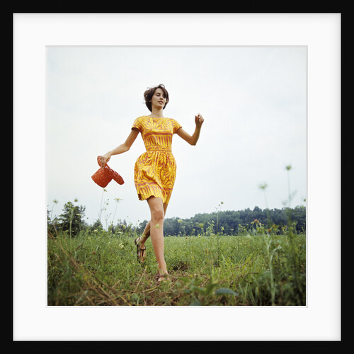 1970s Young Woman Wearing Colorful Print Dress Running In Field Carrying Red Straw Hat by Anonymous