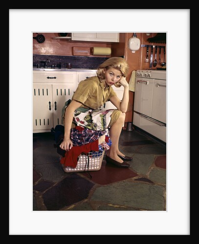 1960s Weary Dejected Woman Housewife Homemaker Sitting On Full Laundry Basket In Kitchen by Anonymous