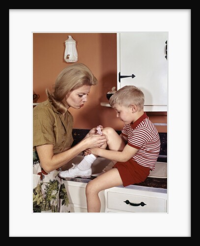1970s Mother Giving First-Aid Putting Band-Aid On Knee Of Son In Kitchen by Anonymous