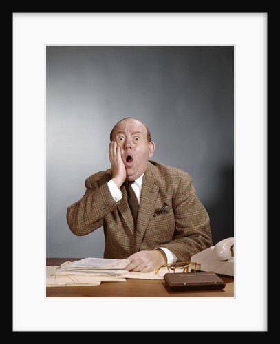 1960s Shocked Balding Executive With Hand To Cheek Sitting At Desk by Anonymous