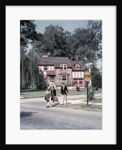 1950s Kids Carry Book Bags Crossing Street By Yellow Stop Sign In Suburban Neighborhood by Anonymous