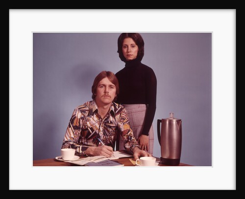 1970s Husband Wearing Loud Print Shirt Writing Family Budget and Wife Standing Behind Coffee Pot by Anonymous