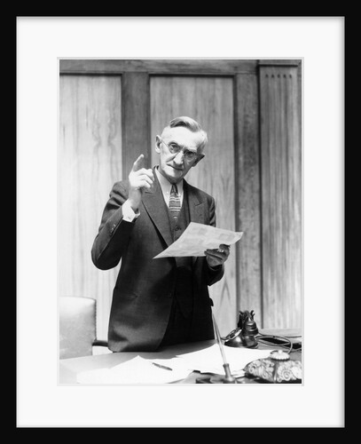 1930s Elderly Man In Office Standing Behind Desk Gesturing With Raised Finger by Anonymous