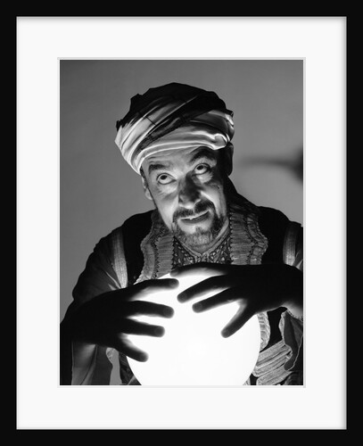 Scary fortune teller man with hands on lighted crystal ball wearing turban by Anonymous