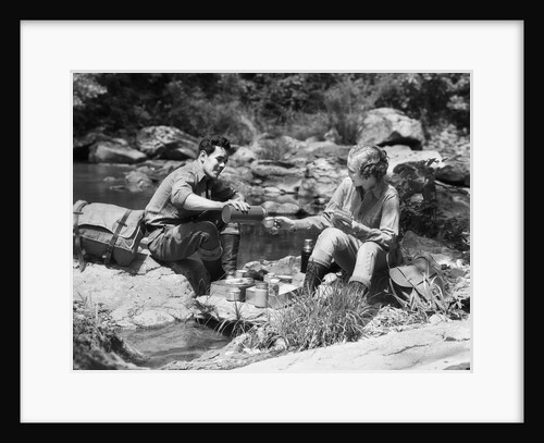 Couple having picnic sitting on rocks by stream man pouring drink for woman from thermos by Anonymous