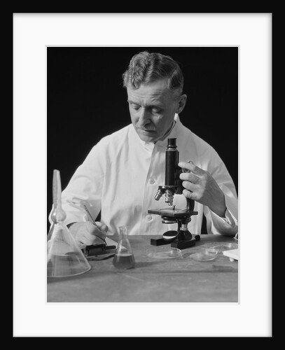 Male scientist wearing white lab coat writing data on clip board holding focus control of microscope by Anonymous