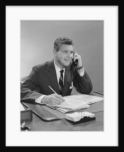 Businessman seated at desk talking on telephone smiling writing with pencil by Anonymous